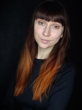 Na czarnym tle ubrana na czarno dziewczyna w ujęciu portretowym. Ma rude włosy, grzywkę, uśmiecha się.