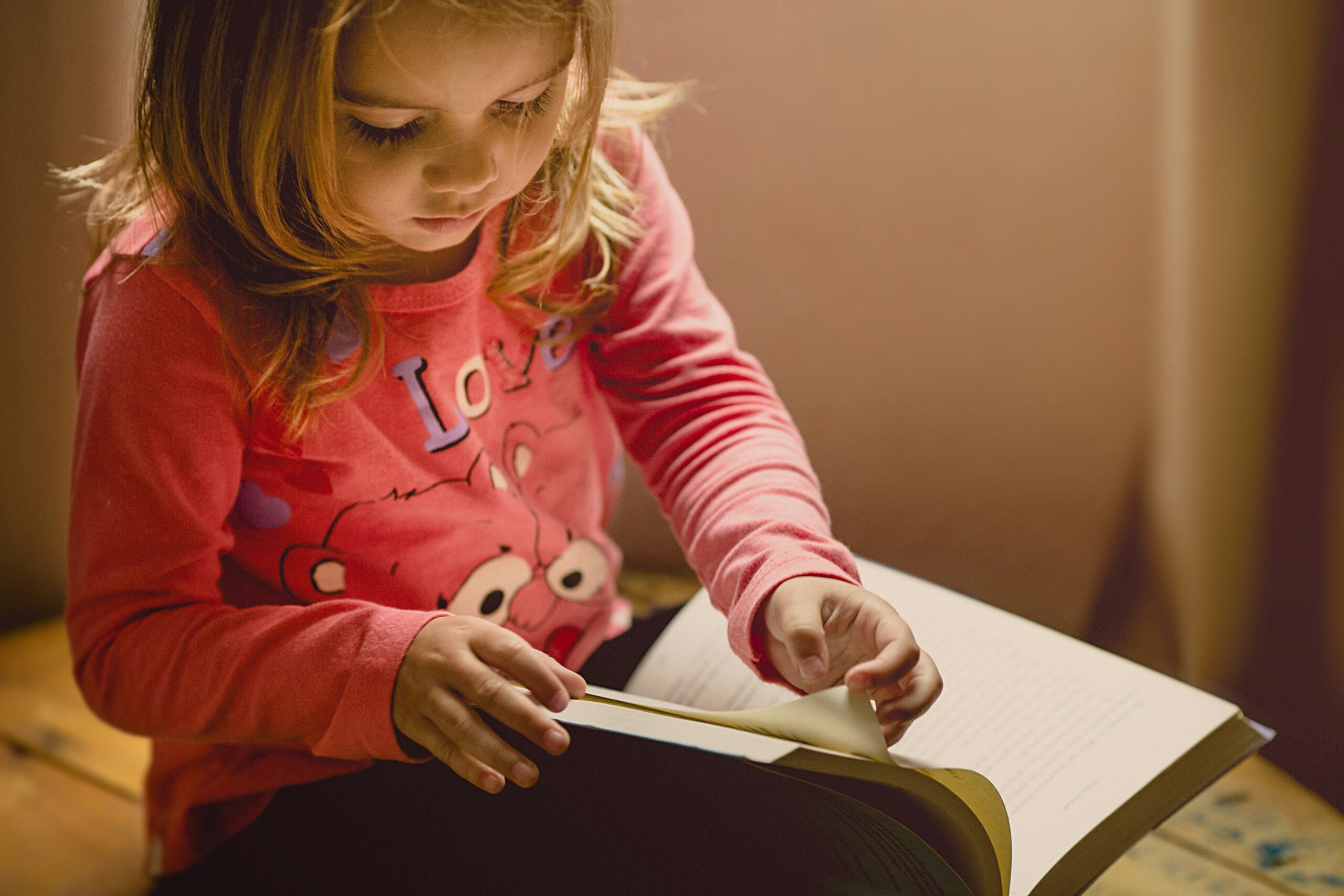 Po lewej stronie kadru widoczna siedząca mała dziewczynka. Na kolanach trzyma rozłożoną książkę. Dziewczynka ma blond włosy do ramion, ubrana jest w różową bluzę we wzorki.