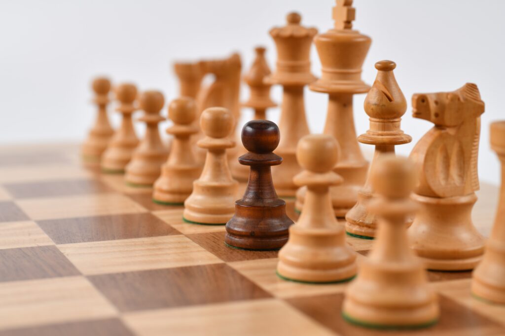 Brązowa szachownica. Z prawej strony ustawione jasne figury szachowe, a w środku pomiędzy nimi jeden czarny pionek. Tło zdjęcia białe.