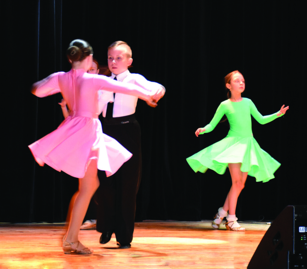 Troje dzieci tańczy na scenie, na tle czarnych kurtyn. Po prawej dziewczyna w zielonej sukience. Po lewej para, dziewczyna w różowej sukience i chłopak ubrany w eleganckie czarne spodnie, białą koszulę i krawat.