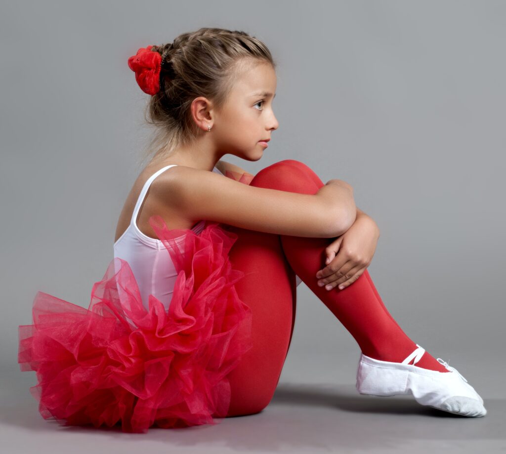 Na szarym tle widoczna z profilu siedząca mała dziewczynka. Ubrana jest w czerwone baletowe tutu, czerwone rajstopy, białe buty i białą koszulkę. W ciemnych włosach ma czerwony kwiatek. Obejmuje kolana rękoma.
