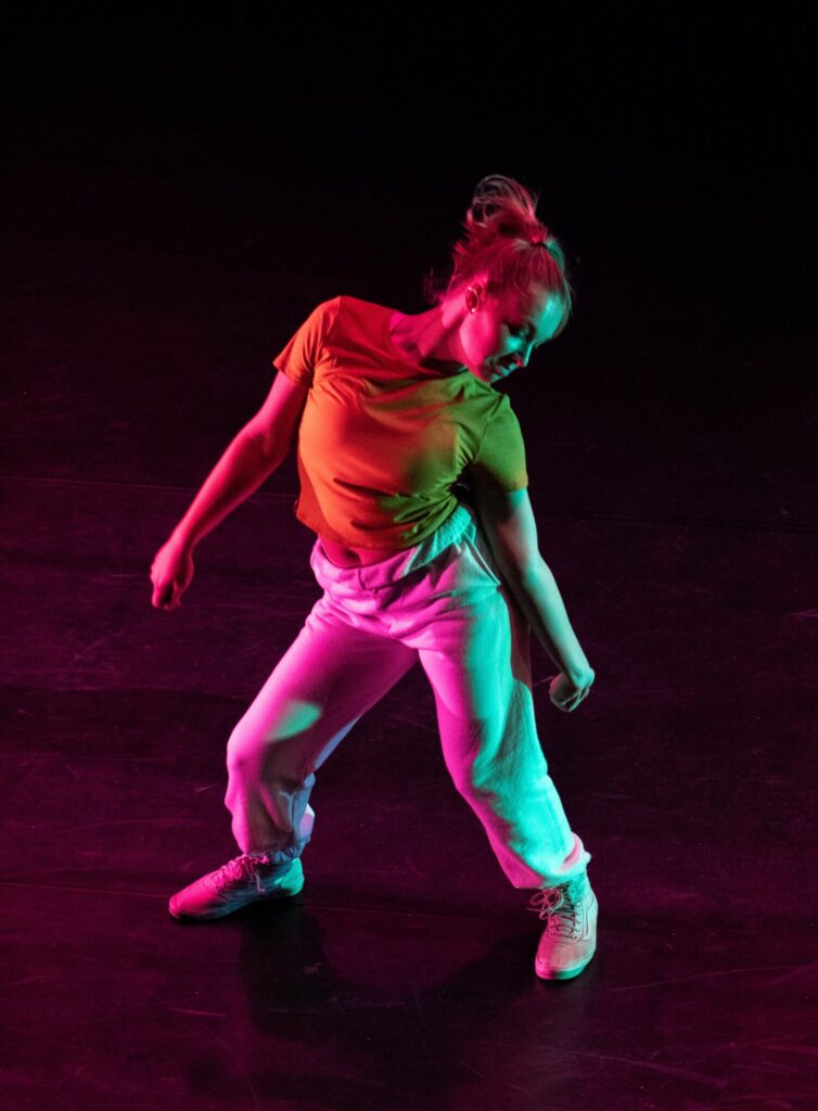 Na ciemnym tle dziewczyna w tanecznej pozie - na szeroko rozstawionych nogach, z głową pochyloną w prawo. Tancerka jest bosa. Oświetlają ją kolorowe światła: różowe, czerwone, zielone i niebieskie.