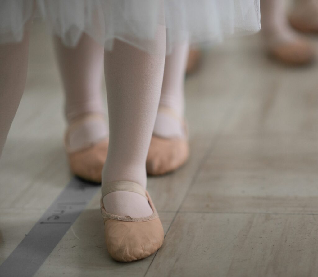 Po lewej stronie zdjęcia widoczne dziecięce nogi w baletkach. W górnym lewym rogu widać także krawędź sukienki baletowej.
