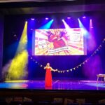 Na oświetlonej na fioletowo scenie, w samym jej centrum, stoi kobieta ubrana w długą czerwoną suknię. W dłoni ma mikrofon, śpiewa piosenkę.