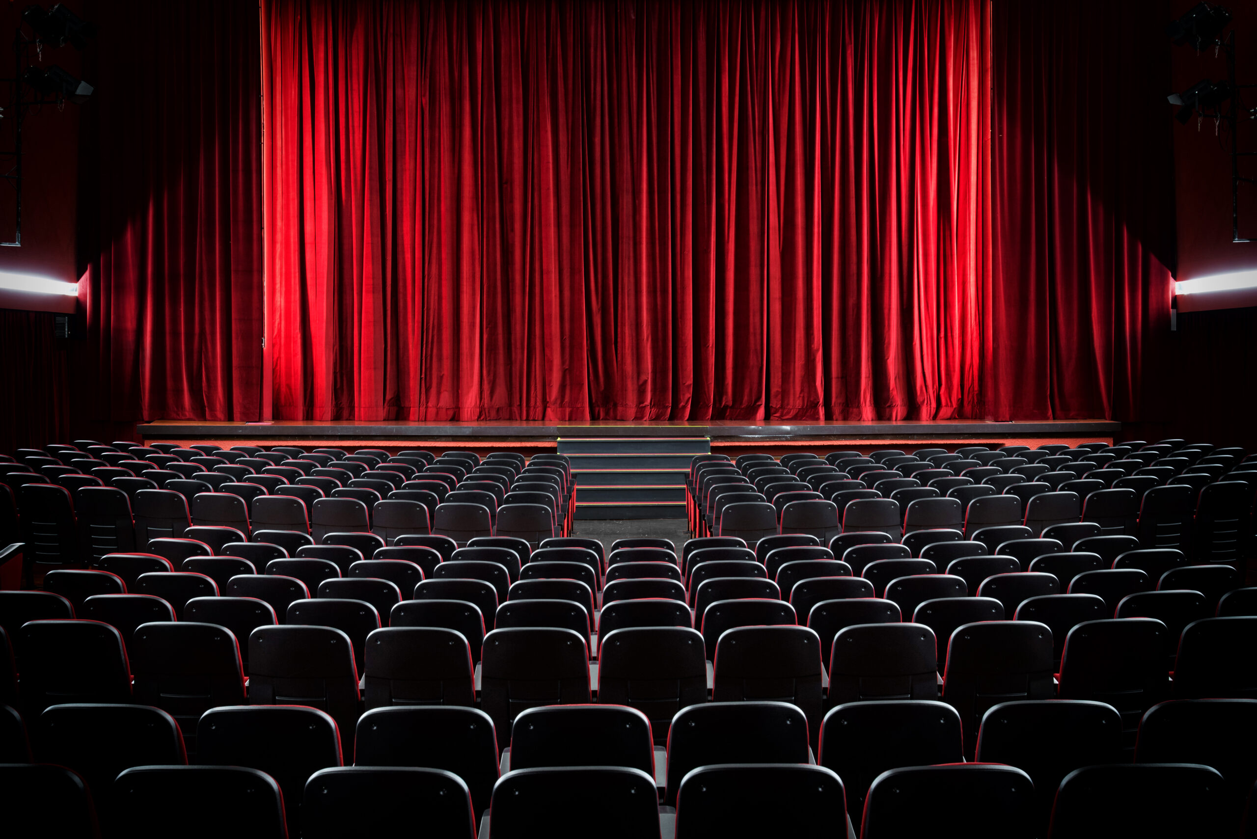 Zdjęcie czerwonej kurtyny w teatrze oraz kilku rzedow foteli na widowni.