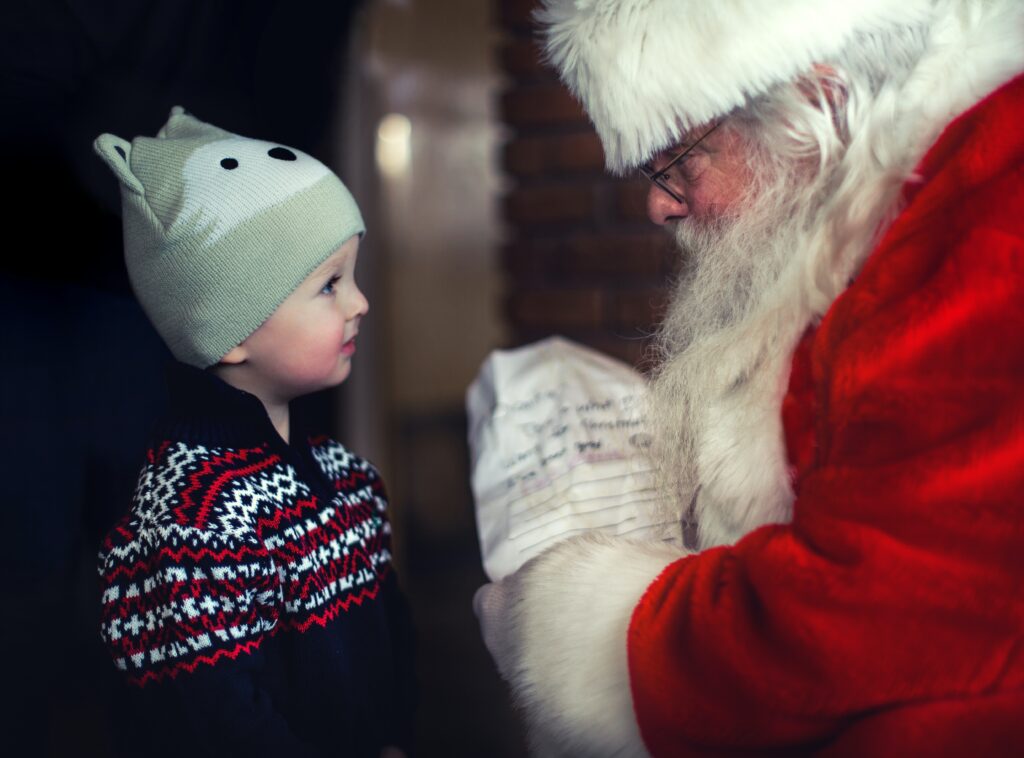 Dwie postaci widoczne z profilu. Po prawej stronie Święty Mikołaj, w czerwonym płaszczu z białym futerkiem, widać brodę, okulary, białe futerko na czapce. W ręku trzyma list. Po lewej stronie małe dziecko, w sweterku w czerwone i granatowe paski i szarej czapeczce z mordką wilka. Dziecko patrzy uważnie na Świętego Mikołaja.