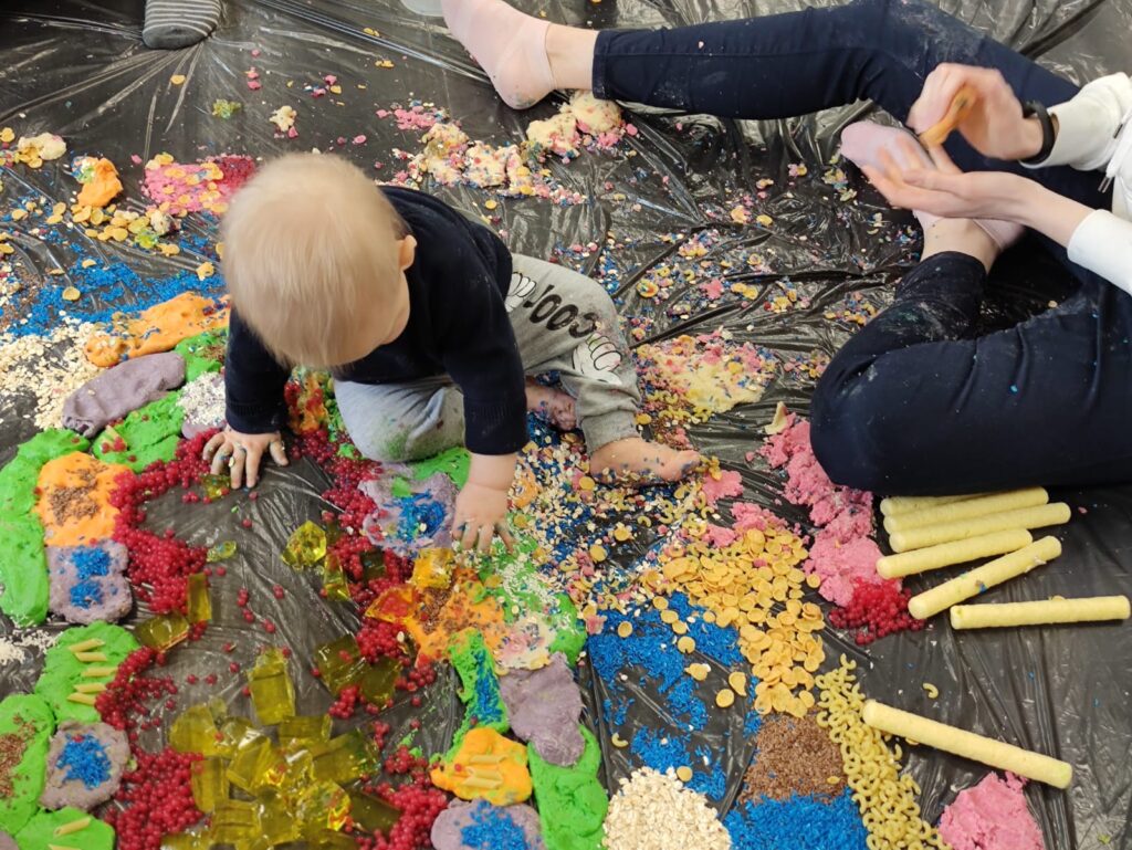Malutkie dziecko bawi się w środku ułożonej z makaronów, mąki, kolorowych proszków, galaretki, chrupek etc mandali. Z boku zdjęcia widać nogi opiekunów, siedzących na podłodze dookoła mandali.