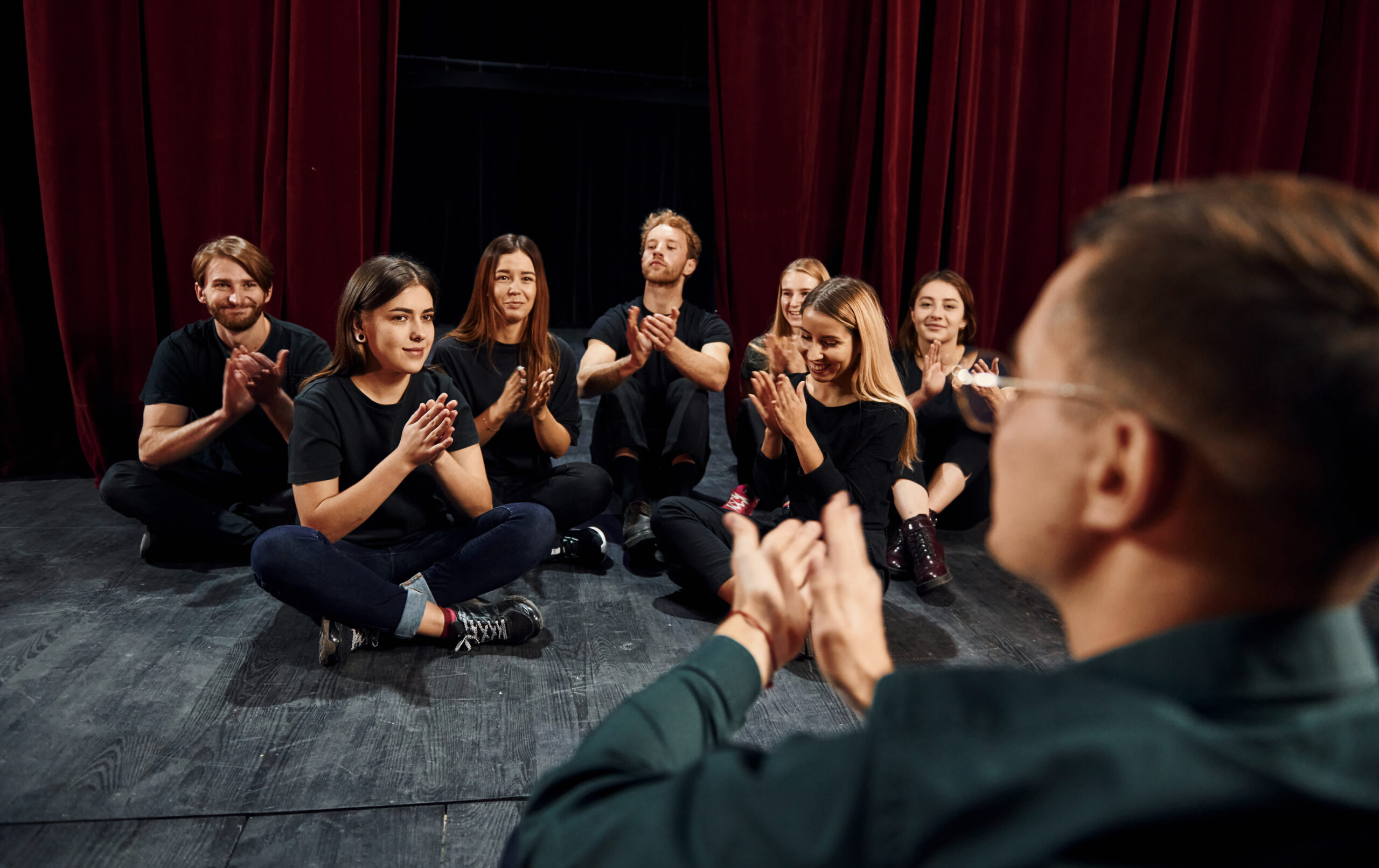 Grupa aktorów siedzi na scenie podczas próby. Wszyscy sa ubrani na czarno i klaszcza w rece podczas cwiczen.