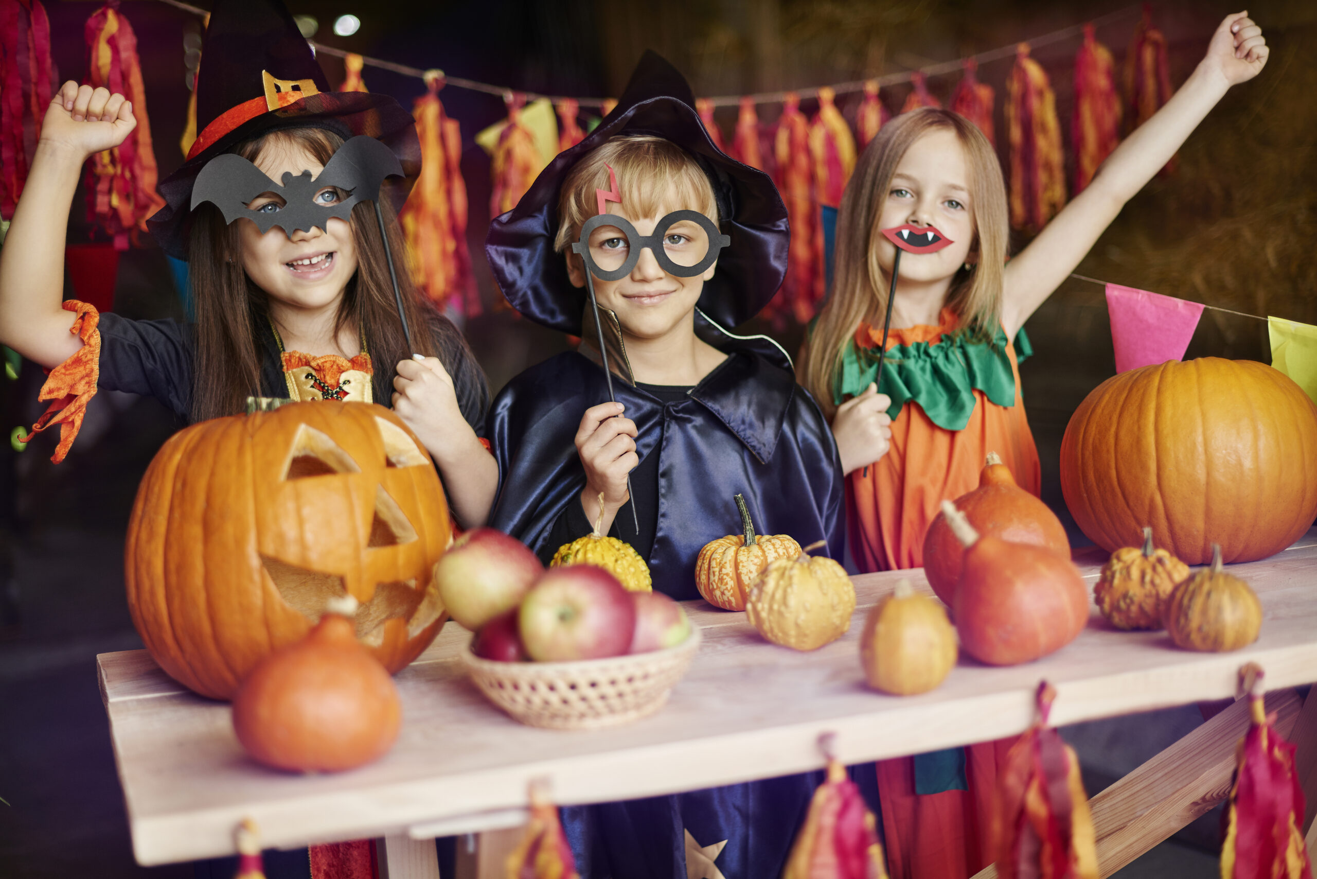 Dwie dziewczynki i chłopiec przebrani za czarodziejów pozują z akcesoriami halloweenowymi, wycietymi dyniami i maskami w ksztalcie nietoperzy.