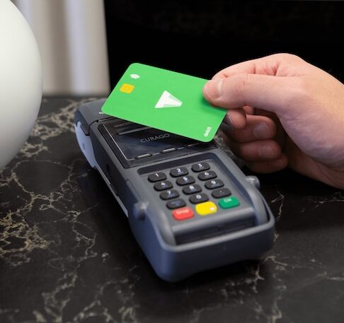 W centrum zdjęcia, na czarnym blacie, terminal do płatności kartą. Z prawej strony widoczna dłoń trzymająca nad terminalem zieloną kartę płatniczą. W tle czarne i białe niewyraźne obiekty.