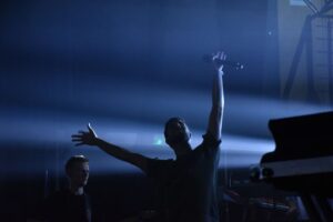 Sylwetka mężczyzny z wyrzuconymi w górę rękami, oświetlona mocnym światłem sceny. Chłopak trzyma w prawej ręce mikrofon.