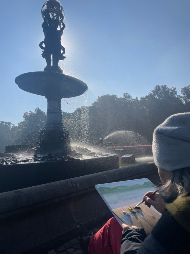 Na zdjęciu kobieta w czapce maluje fontannę przed ktora siedzi.