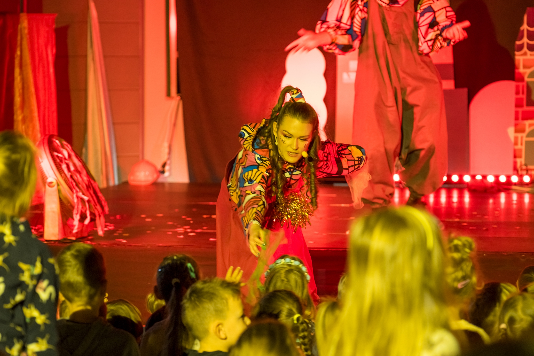 Zdjęcie ze spektaklu, kobieta ubrana na czerwono podaje rękę dzieciom z widowni.