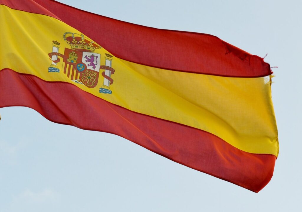 Na tle niebieskiego nieba flaga hiszpanska: czerwone pasy poziome na górze i na dole, pomiędzy szerszy pas żółty, po lewej stronie godło.