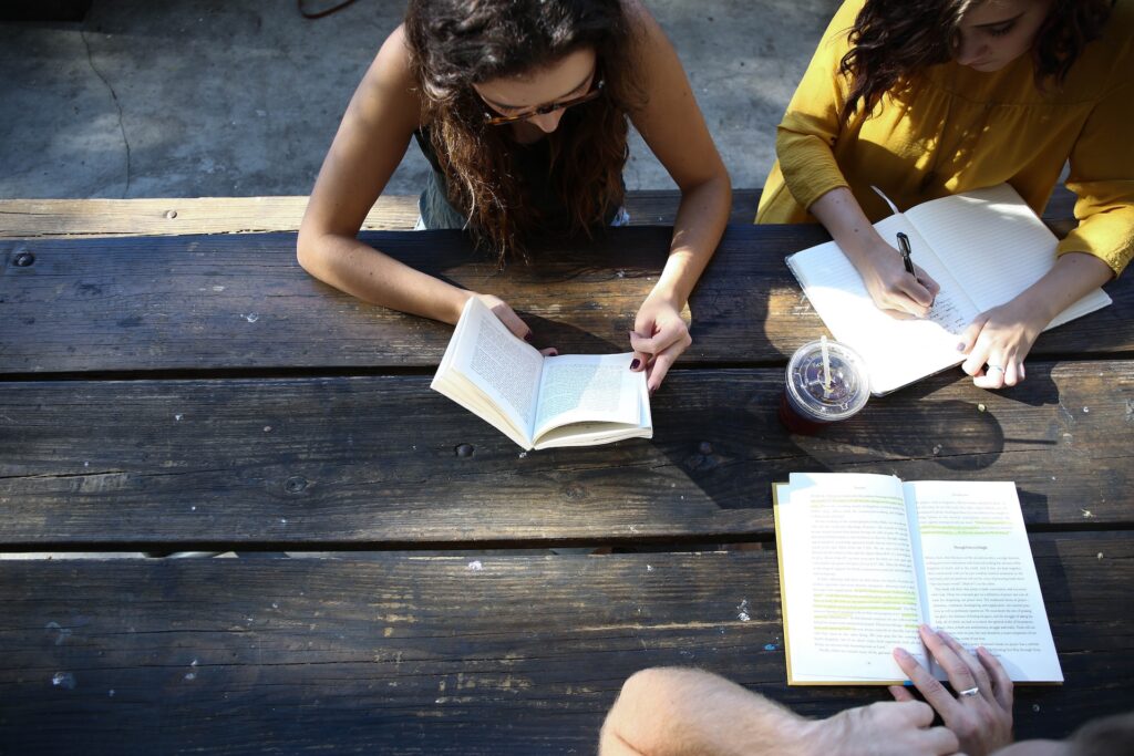 Widok z góry. Na drewnianym szarym stole leżą książki, przy stole trzy dziewczyny uczą się wspólnie.