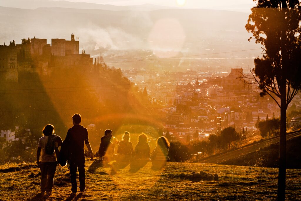Widok hiszpańskiego miasta w zachodzącym słońcu. Na pierwszym planie, na wzgórzu, siedzi trójka ludzi, z lewej strony zdjęcia stoją kolejne dwie postaci. Całość w odcieniach żółci i pomarańczu.