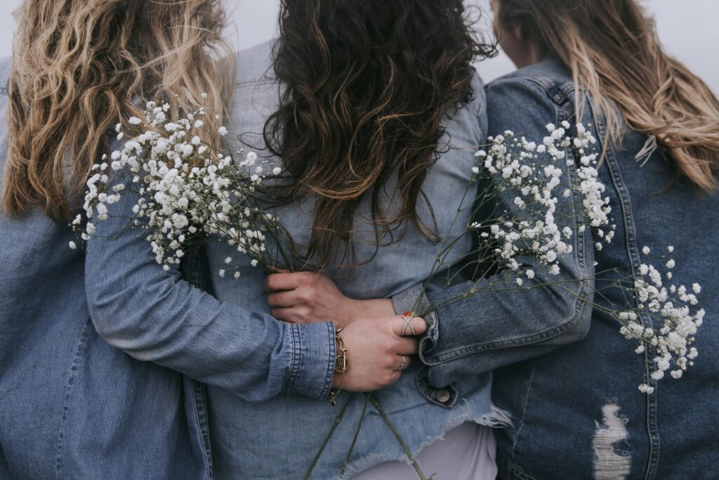 Trzy kobiety stoją tyłem do widza. Są ubrane w niebieskie kurtki, mają długie włosy. Obejmują się przyjacielsko, dwie z nich trzymają w rękach białe kwiaty.