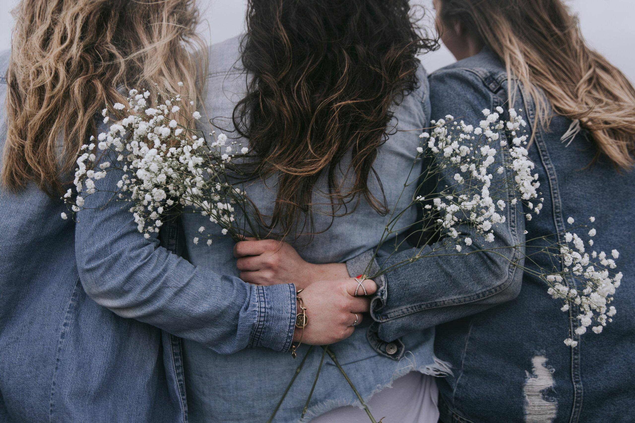 Trzy kobiety stoją tyłem. Są ubrane w niebieskie kurtki, mają długie włosy. Obejmują się przyjacielsko, dwie z nich trzymają w dłoniach białe kwiaty.