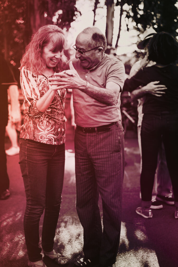 Po lewej stronie na pierwszym planie tańcząca starsza para. Stoją przodem do widza, trzymają się za ręce w tanecznej pozie, głowy mają pochylone ku sobie. Oboje uśmiechają się radośnie. Zdjęcie ma zmodyfikowane kolory - nałożony czerwono-fioletowy filtr.