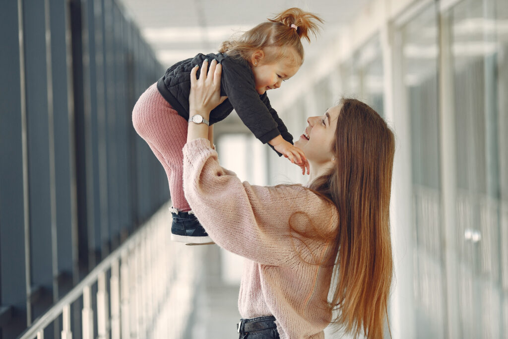 Zdjęcie kobiety podnoszącej do góry małą dziewczynkę. Kobieta uśmiecha się do dziecka, dziewczynka wyciąga do niej rączki.
