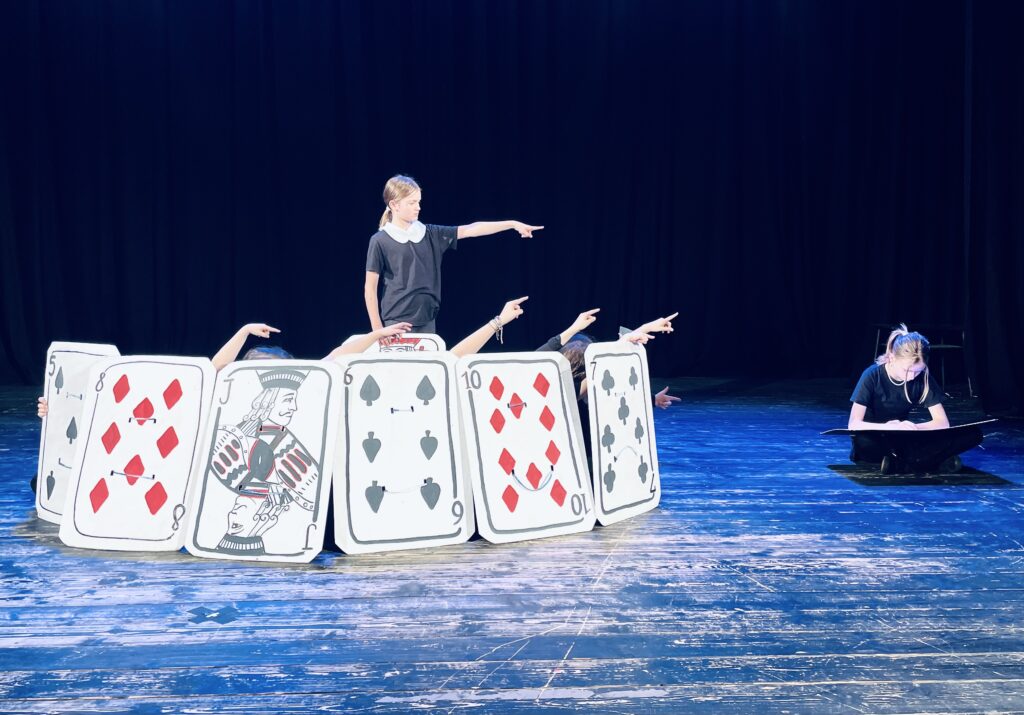 Zdjęcie ze spektaklu, dziewczynka stoi w kręgu ułożonym z wielkich kart do gry i wkazuje palcem na dziecko siedzące poza kręgiem.