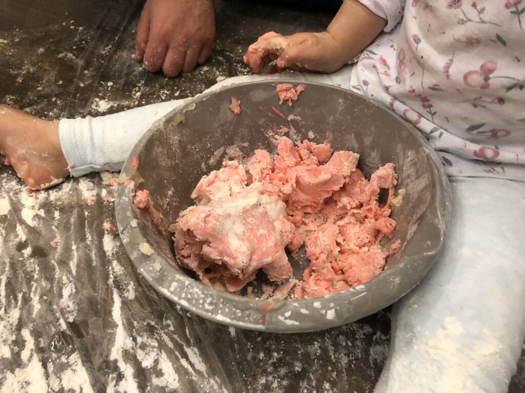 Szara plastikowa miska wypełniona jest czerwoną masą. Przy misce okrakiem siedzi dziecko, widać jego nogi w białych getrach i dłoń ubrudzoną masą z miski.