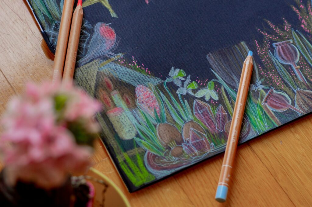 Na drewnianym blacie leży czarny papier, widoczny w górnej prawej części zdjęcia. Na papierze leżą kredki. Narysowany obrazek: wiosenne kwiaty, ptak z czerwonym brzuszkiem.