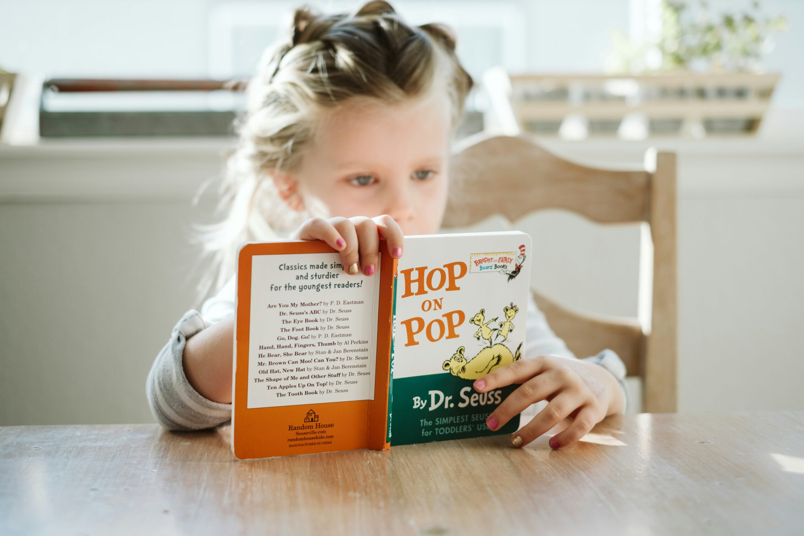 Przy drewnianym stole mała dziewczynka czyta książkę. Opiera ją pionowo o blat. Książka ma zielono-pomarańczową okładkę.