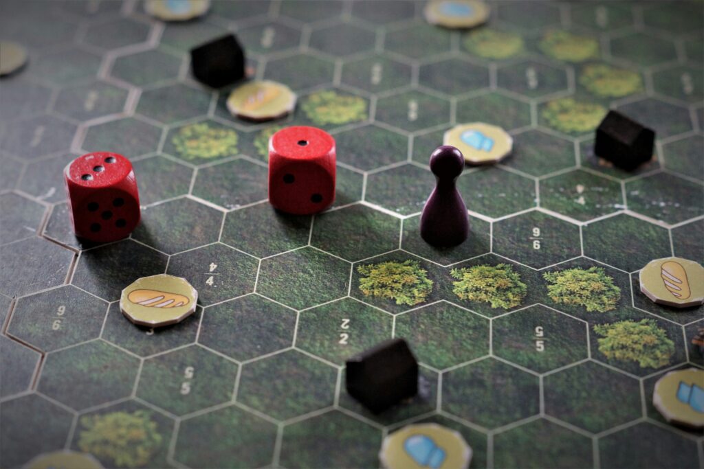 Na zielonej planszy do gry stoją czarne pionki, leżą złote monetki i czerwone kości do gry.