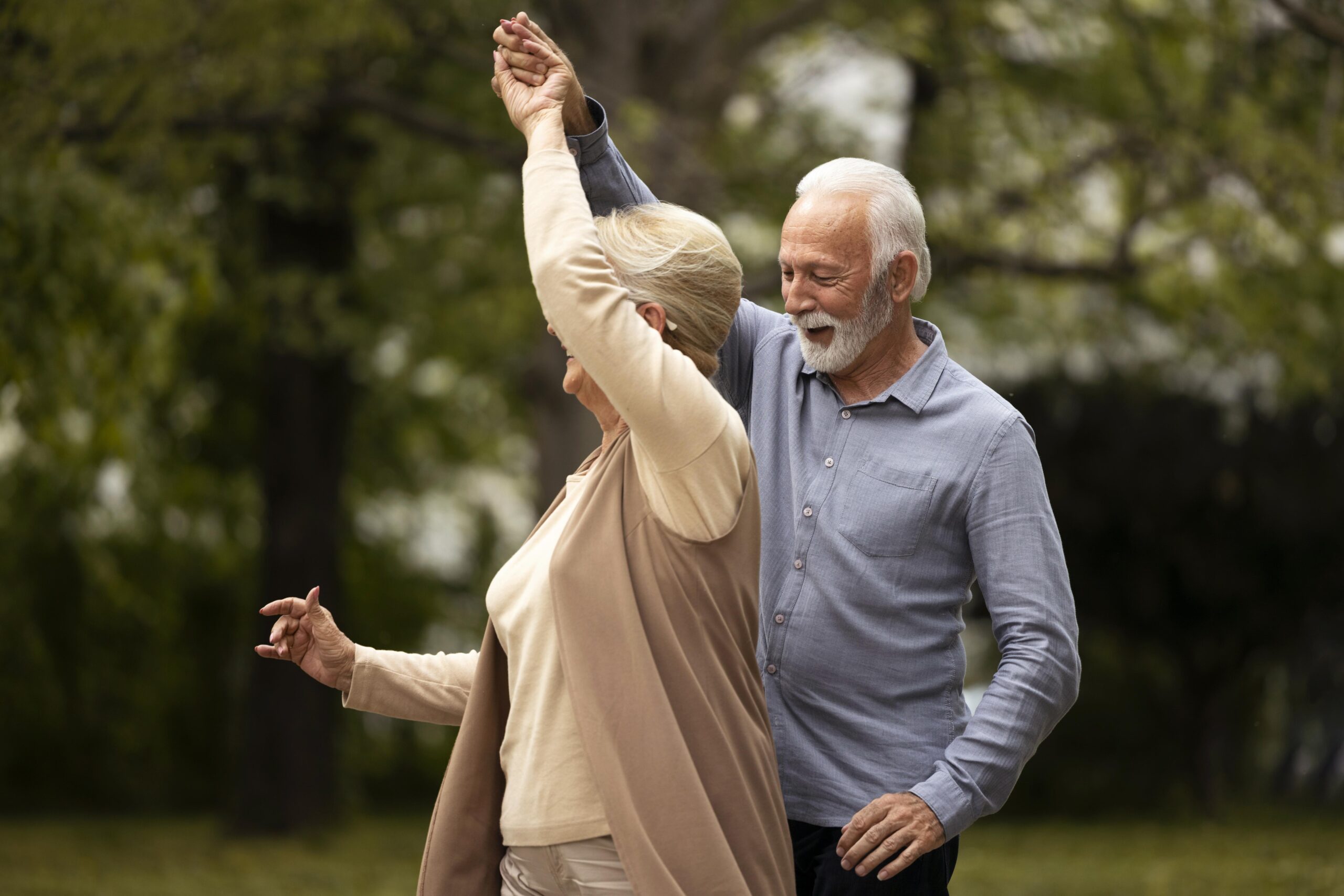 Para seniorów tańczy w parku. Mężczyzna ubrany jest w niebieską koszulę, kobieta w beżowy sweterek. Mężczyzna, unosząc dłoń, prowadzi kobietę do piruetu.
