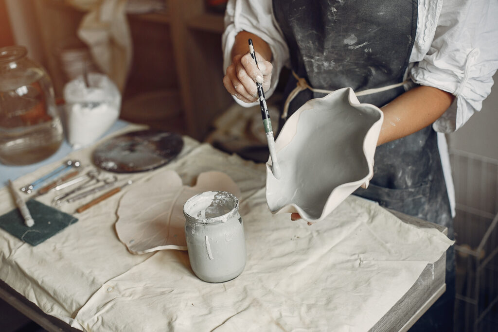 Zbliżenie na kobiece ręce trzymające talerz. Kobieta w pracowni ceramicznej maluje go szarym szkliwem.