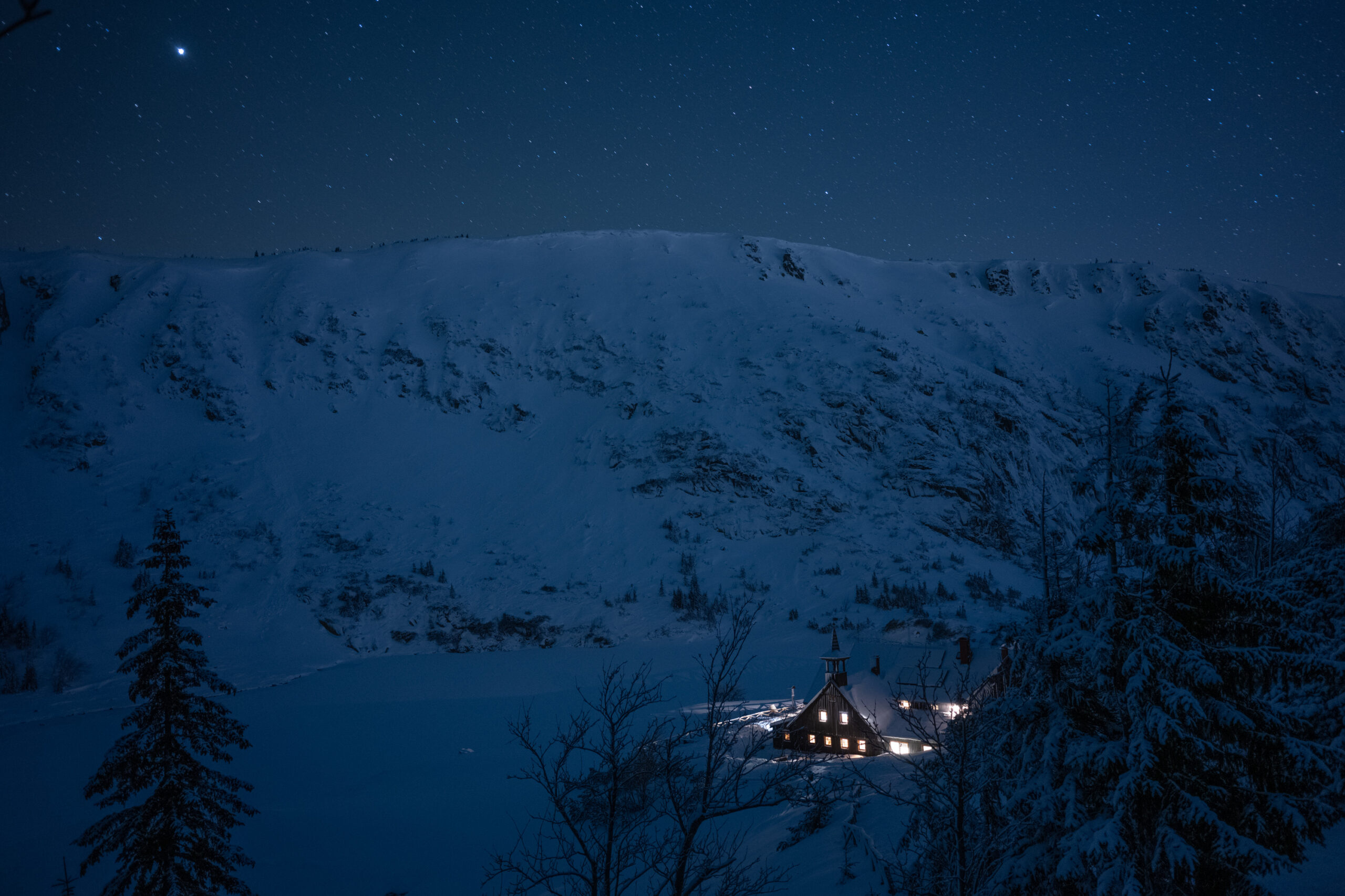 Zdjęcie przedstawia zimowy, nocny, górki krajobraz. W dolinie znajduje się rozświetlony schronisko Samotnia.