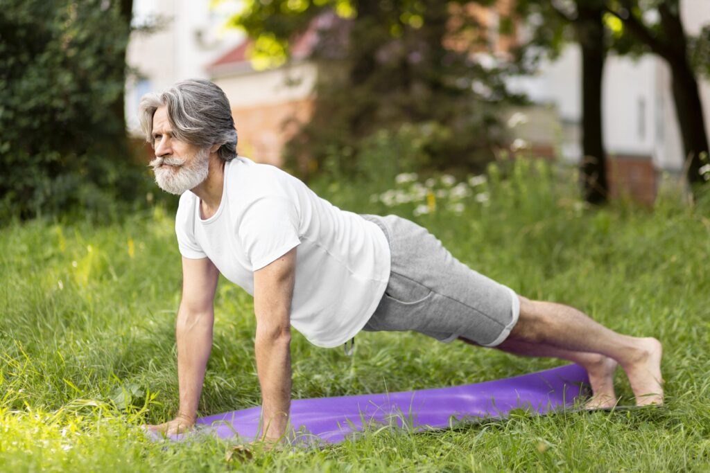 Starszy brodaty mężczyzna ćwiczy jogę na trawie. Na fioletowej macie przyjmuje pozycję podporu na rękach i stopach.