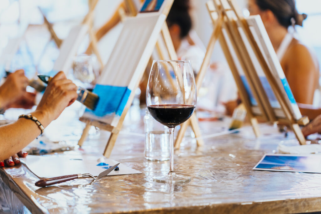 Zbliżenie na dłonie osób malujących obrazy na warsztatach. Na stole są ustawione kieliszki z czerwonym winem.