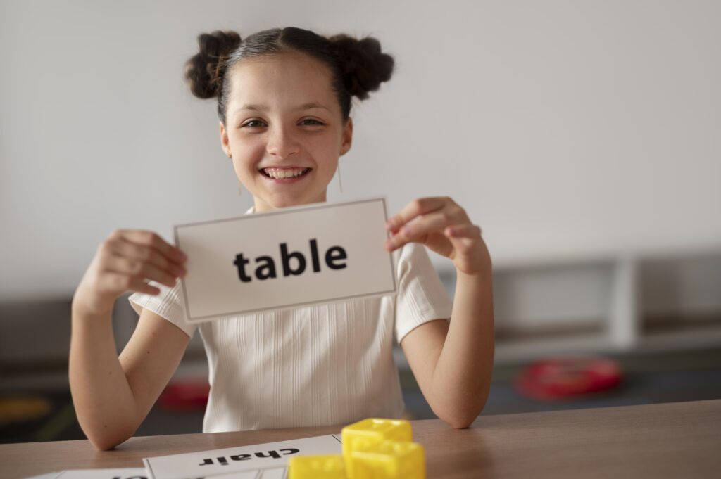 Dziewczynka trzyma oburącz tabliczkę z napisem "table" (stół po angielsku), uśmiecha się. Ubrana jest w białą bluzkę, a jej ciemne włosy są spięte w dwa koki po obu stronach głowy, blisko przy uszach.