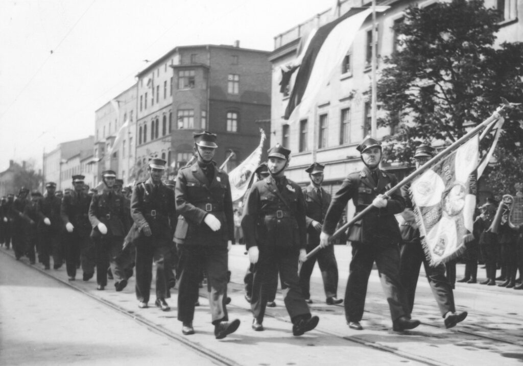 Archiwalne zdjęcie przedstawiające defiladę związku powstańców śląskich z 1938 roku. Na czele idzie poczet sztandarowy. Zdjęcie wykonano na skrzyżowaniu ulicy 1 Maja z ulicą Bogucicką.