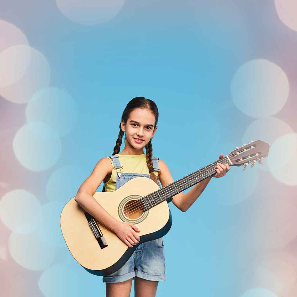 Na niebieskim tle stoi dziewczynka z gitarą. Ma czarne warkocze, ubrana jest w żółtą koszulkę i niebieskie jeansy.