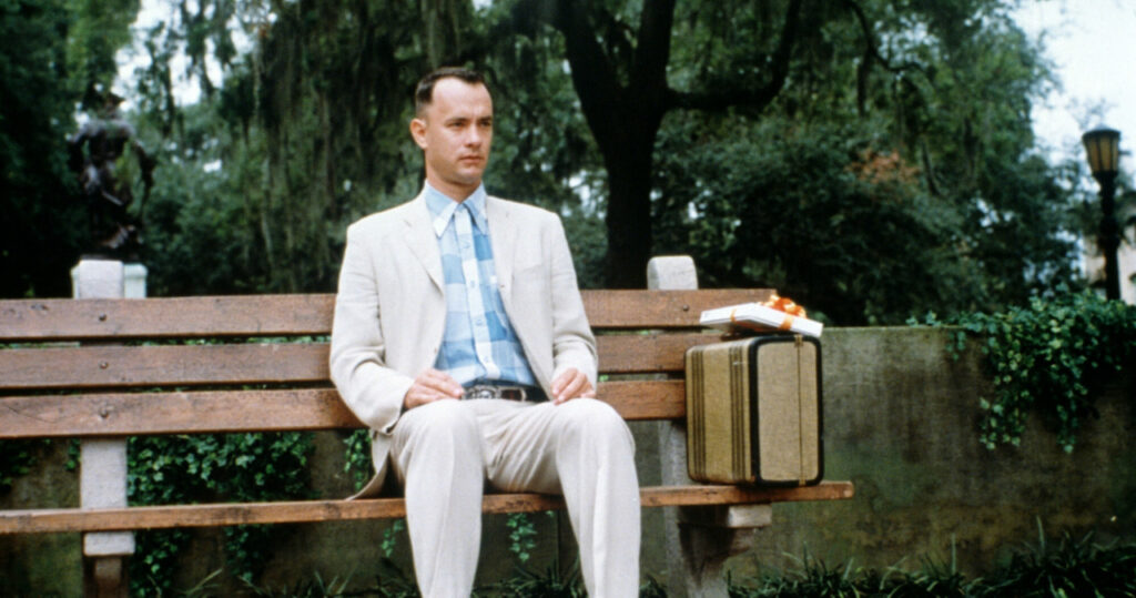Kadr przedstawiający scene z filmu. Forrest Gump (brunet w białym garniturze) siedzi na ławce z postawioną obok walizką.