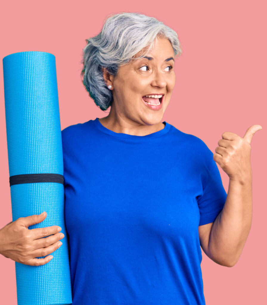 Na jednolitym różowym tle stoi uśmiechnięta siwa kobieta w niebieskiej koszulce. W prawej ręce trzyma niebieską matę do jogi. Lewą ręką pokazuje znak ok - kciuk do góry.