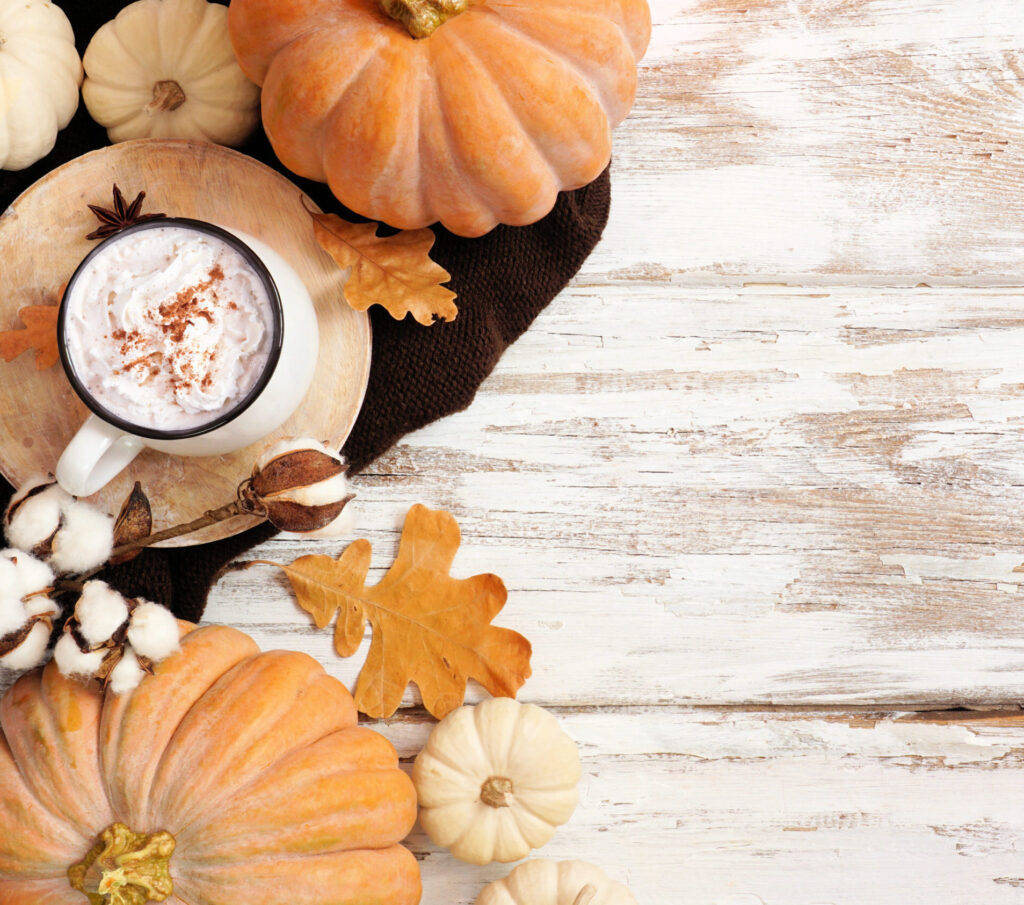 Widok z góry. Blat drewnianego stołu przecieranego na biało. Z lewej strony na stole kompozycja dyń i jesiennych liści, a pomiędzy nimi na brązowej tacce kubek kawy z mleczną pianką.