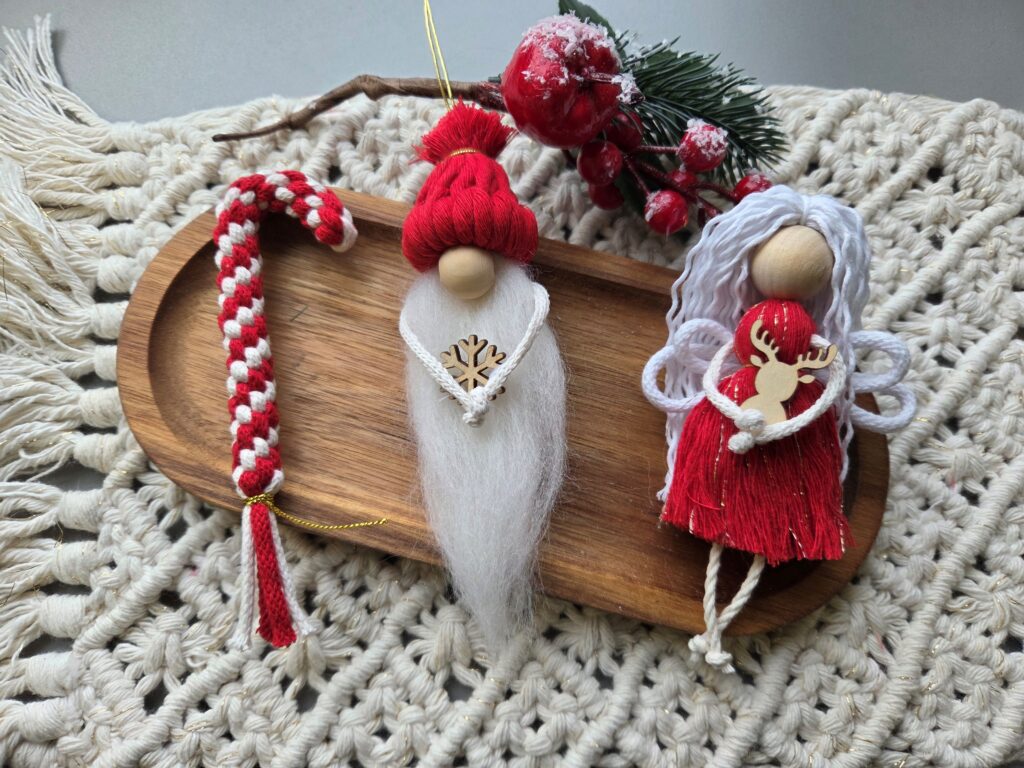 Fotografia, widok z góry: na makramowym obrusie położona drewniana patera, a na niej trzy świąteczne makramowe ozdoby: aniołek w czerwonej sukience, mikołajek składający się z czerwonej czapki i białej brody, biało-czerwona laseczka-cukierek. Za ozdobami dekoracyjna zielona gałązka z czerwonymi owockami.