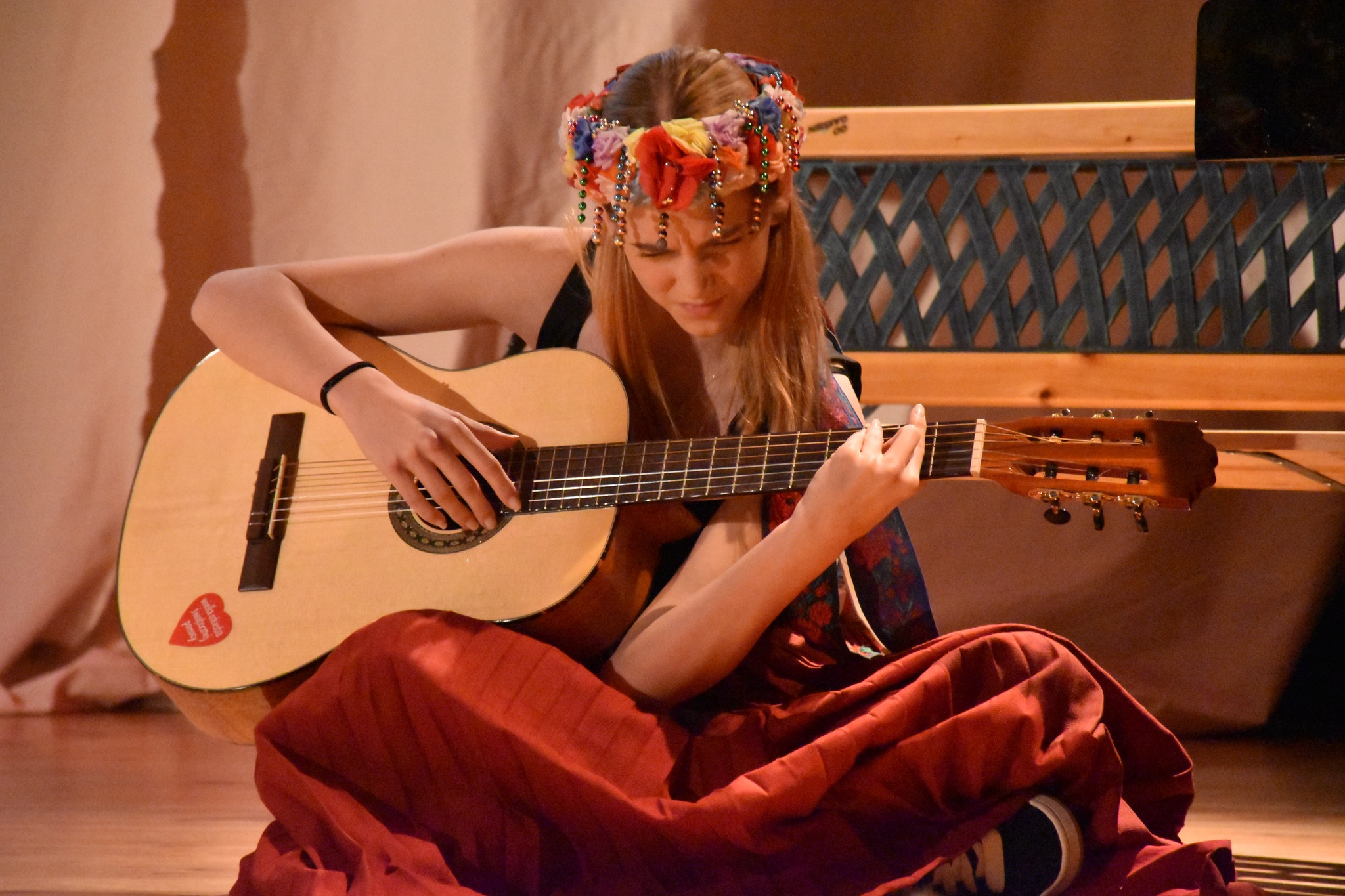 Fotografia ze spektaklu: na środku po turecku siedzi długowłosa dziewczyna w czerwonej spódnicy i śląskim wianku. Gra na gitarze, oparetj o kolano. W tle za nią drewniana ławka.