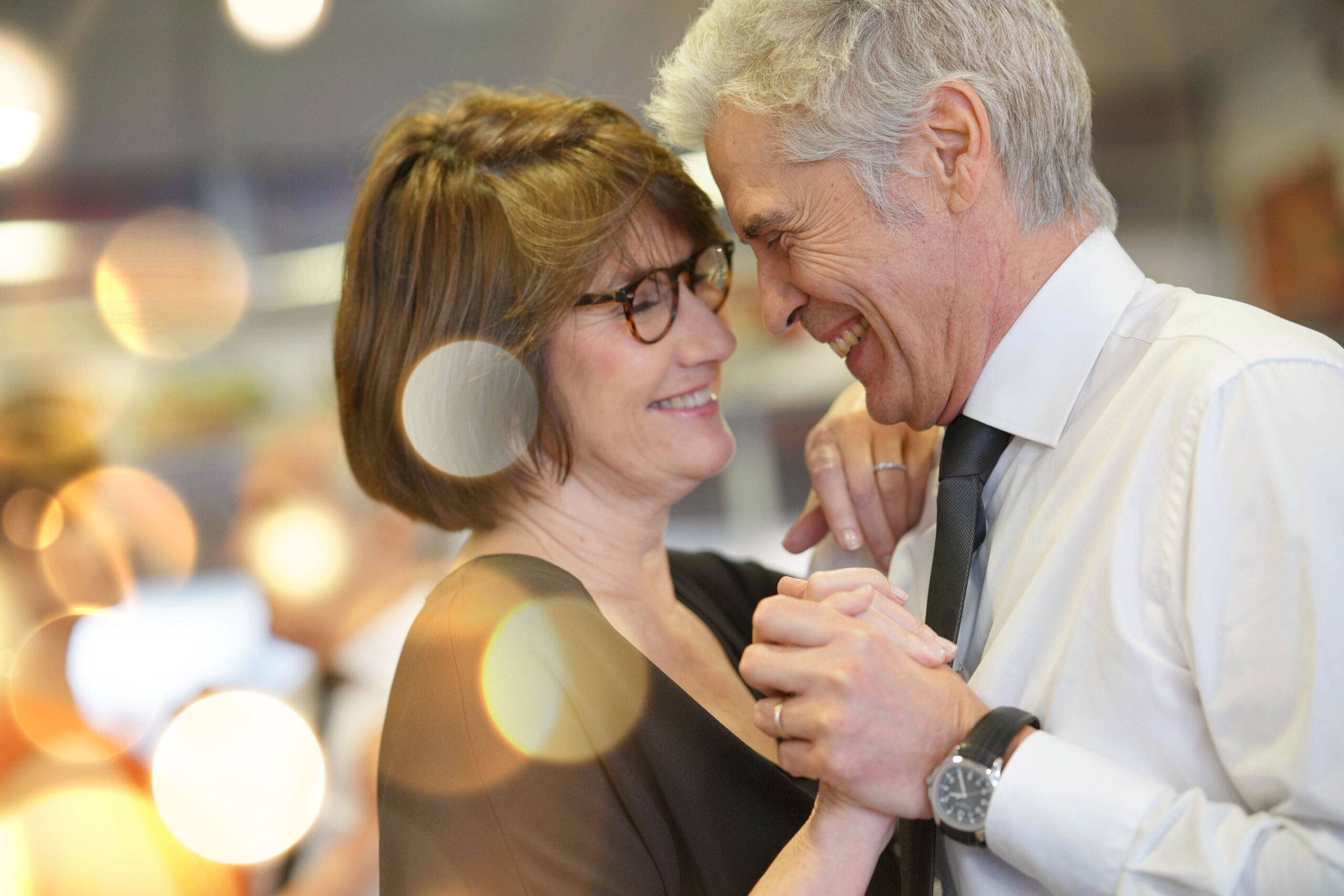 Starsza kobieta i starszy mężczyzna tańczą razem przytuleni, spoglądając na siebie z uśmiechem. Są widoczni w zbliżeniu od piersi w górę, on ma elegancką koszulę i krawat, ona czarną sukienkę. W tle rozmazane światełka.