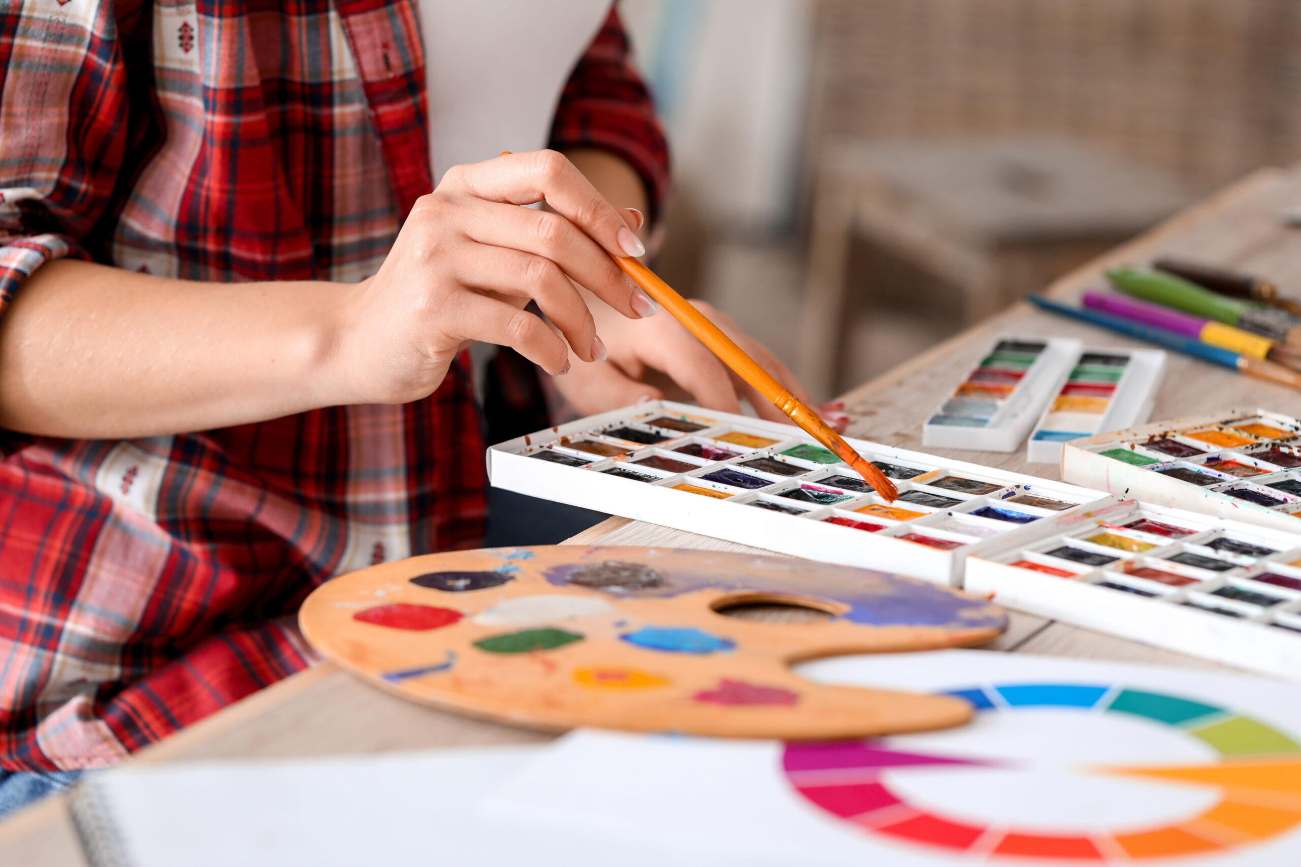 Widoczny fragment stołu z rozłożonymi farbami akwarelowymi, drewnianą paletą, kartką z kołem barw, kartką z próbkami kolorów. Z lewej strony osoba w czerwonej koszuli w kratę, widać jej dłoń z pędzlem.