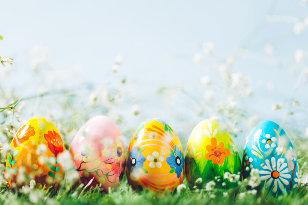 Zmodyfikowane zdjęcie: na dole na pierwszym planie rząd kolorowych malowanych jajeczek ustawionych w trawie i ozdobionych białymi kwiatuszkami. Za nimi rozmazane niebieskie tło z zarysami listków i bazi.