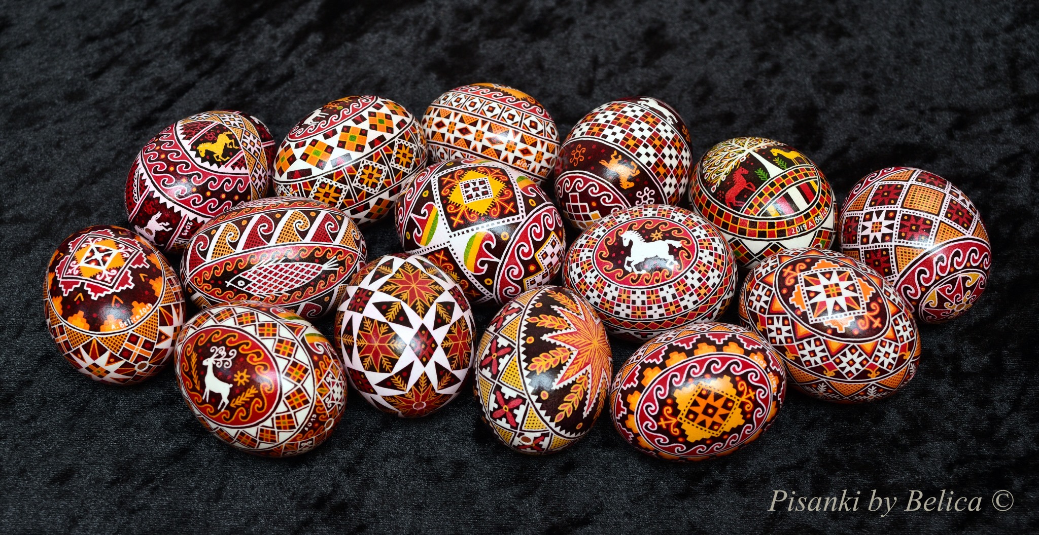 Na ciemnym tle leży dużo pisanek ozdobionych precyzyjnymi wzorami w odcieniach brązu, żółci, czerwieni i pomarańczu.
