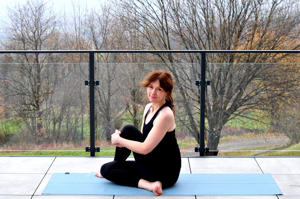 Na niebieskiej macie siedzi ubrana na czarno jasnowłosa kobieta: Ania Gościniewicz, instruktorka jogi. Za nią widoczna przeszkolona balustrada balkonu, a w tle bezlistne drzewa.