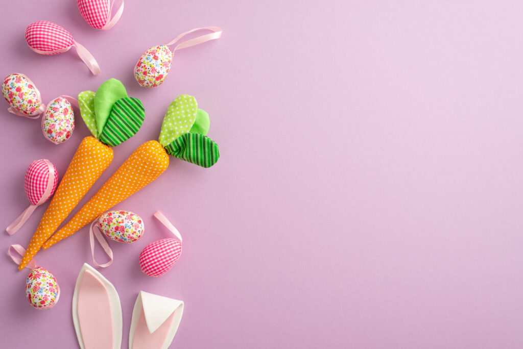 Widok z góry. Na różowym tle rozłożone ozdoby uszyte z materiałów: pomarańczowe marchewki, białe i różowe jajeczka z kokardką, królicze uszka z filcu.