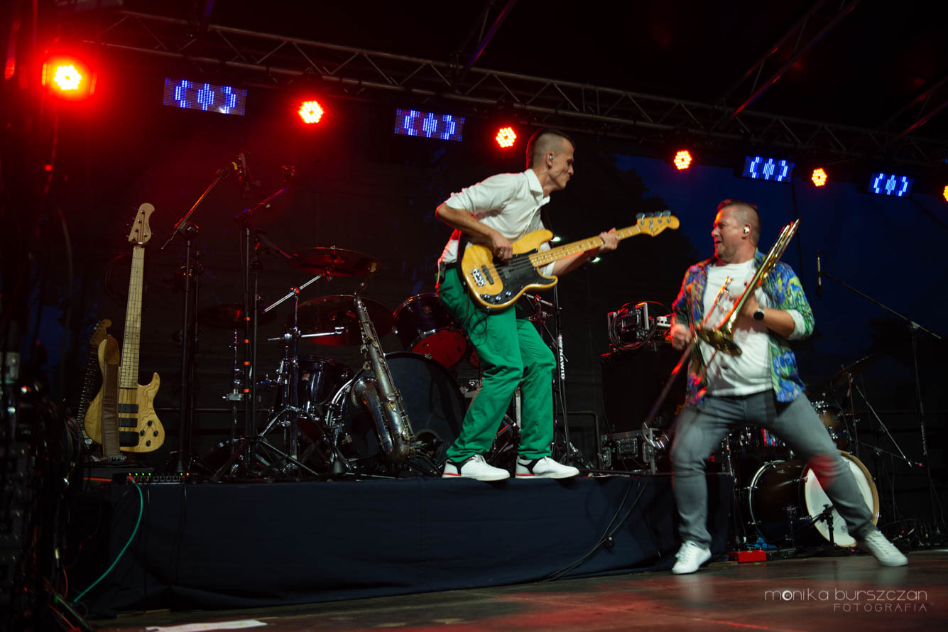 Fotografia z koncertu - na kolorowo oświetlonej scenie dwóch muzyków - na podwyższeniu mężczyzna w białej koszuli i zielonych spodniach, z żółtą gitarą, na scenie gitarzysta w dżinsach i dżinsowej koszuli.